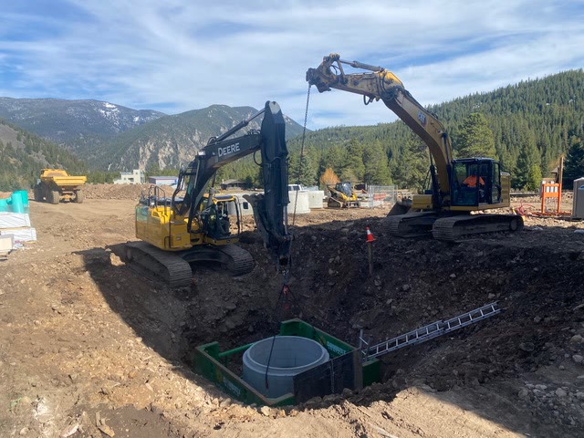 Two excavators set a concrete vault into a shored excavation with mountains behind